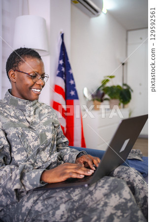 Smiling african american female soldier using laptop at home with United States flag in background 129592561