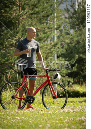Cyclist wearing gray t-shirt stand near bicycle at greenery public park. Summer outdoor activity. Vertical format 129593587