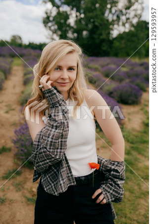Blonde woman touching her face while posing in lavender field, wearing checkered shirt over white tank top and black trousers, with red poppy flower in her belt. Portrait of blonde woman touching face Blonde woman touching her face while posing in lavender field, wearing checkered shirt over white tank top and black trousers, with red poppy flower in her belt. Portrait of blonde woman touching face 129593957