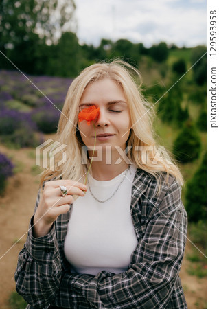 Young blonde woman with closed eyes holding red poppy flower in front of her eye, enjoying scent and atmosphere in lavender field, wearing casual clothing. Blonde woman holding red poppy close to face Young blonde woman with closed eyes holding red poppy flower in front of her eye, enjoying scent and atmosphere in lavender field, wearing casual clothing. Blonde woman holding red poppy close to face 129593958