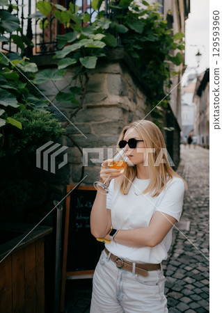 Young adult blonde woman enjoying glass of orange wine in picturesque European cobblestone alley, surrounded by charming stone buildings and lush greenery. Caucasian woman sipping orange wine Young adult blonde woman enjoying glass of orange wine in picturesque European cobblestone alley, surrounded by charming stone buildings and lush greenery. Caucasian woman sipping orange wine 129593960