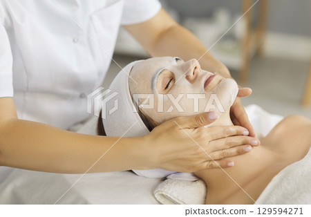 Young woman in spa salon receiving beauty procedure with moisturizing mask from beautician 129594271