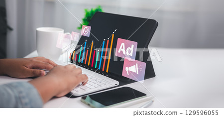 Professional reviewing digital marketing analytics on a tablet with rising bar graph, ad icons, and e-commerce symbols, representing online advertising success and business growth. 129596055