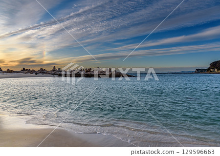 Scenic coastal view with calm sea, sandy beach, and rock formations under a vibrant sky at sunset 129596663