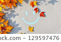 Autumn leaves in warm tones frame chalk smiley, autumn shadows stretch softly across surface, autumn mood glows through playful leaf placement and cheerful seasonal message, copy space 129597476