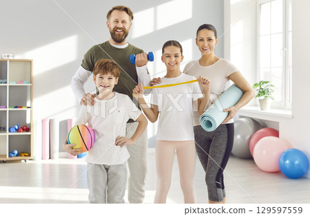 Sporty family with kids standing in gym with sports equipment in hands and looking at camera Sporty family with kids standing in gym with sports equipment in hands and looking at camera 129597559