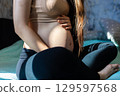 A pregnant woman sits on a bed, gently stroking her belly, enjoying the excitement and anticipation of motherhood, preparing for childbirth. The sun's rays fall on the pregnant woman's belly. 129597568