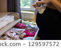 Pregnant woman holding small white baby shoes while organizing and sorting baby clothes, eagerly preparing for the joyful arrival of her newborn and embracing motherhood. 129597572