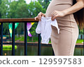 Pregnant woman holding small white baby clothes in her hands, standing on a balcony with colorful flags and a pond in the background, celebrating upcoming motherhood. 129597583