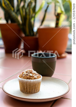 cup of coffee and muffin sit on white plate on pink tiled table. green plants in pots on table. 129597609