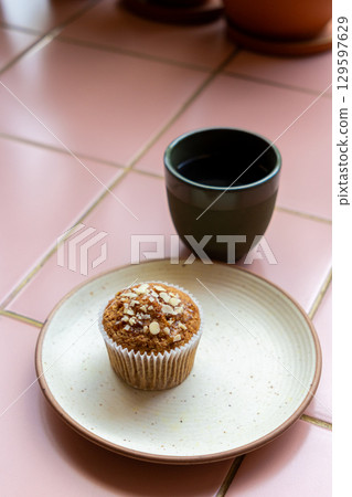cup of coffee and muffin sit on a white plate on pink tiled table. muffin is sprinkled with nuts. 129597629