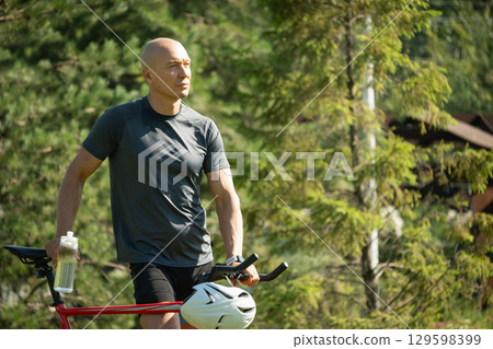 Caucasian cyclist tired, and relax near bicycle at greenery public park. Relaxation and refreshment outdoors among greenery. Take a break. Summer activity. Caucasian cyclist tired, and relax near bicycle at greenery public park. Relaxation and refreshment outdoors among greenery. Take a break. Summer activity. 129598399