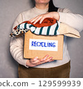 Woman holding cardboard box full of clothes labeled Recycling. Concept of sustainable fashion and responsible wardrobe management. 129599939