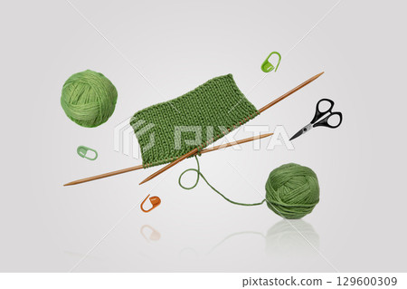 Tools for knitting warm clothes: scissors, green ball of thread, stoppers, and knitting needles with a knitted green piece of fabric flying in the air on a gray background. Hobby home crafts. 129600309