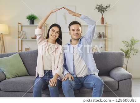 Happy couple celebrating relocation making hand roof above heads sitting on sofa in living room. Happy couple celebrating relocation making hand roof above heads sitting on sofa in living room. 129600385