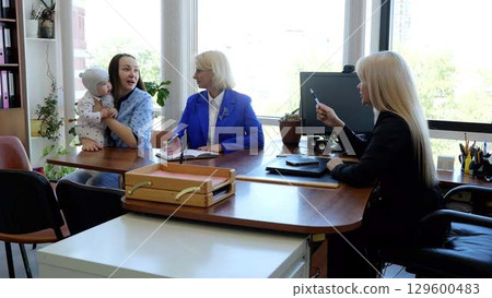 Three female professionals in a contemporary office setting, one multitasking with a baby on her lap 129600483