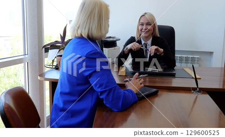 Two businesswomen are sitting at a desk in a modern office, engaged in a discussion, possibly strategizing or problem solving, with a laptop and other office supplies visible on the desk 129600525