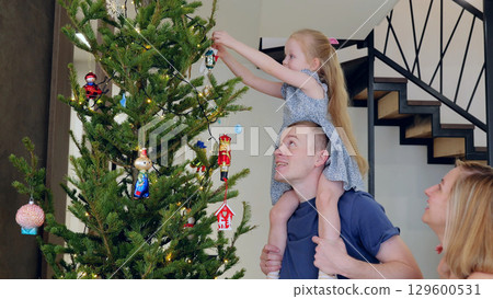 Young family decorates a christmas tree in their modern home, enjoying the festive holiday atmosphere. Daughter decorates a Christmas tree while sitting on her dad's neck Young family decorates a christmas tree in their modern home, enjoying the festive holiday atmosphere. Daughter decorates a Christmas tree while sitting on her dad's neck 129600531