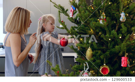 Mother and daughter joyfully decorate a christmas tree, bonding over festive ornaments and twinkling lights in their cozy home. New Year's Eve with family 129600533