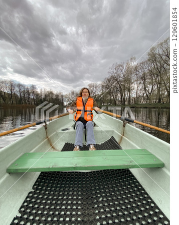 A woman in an orange life vest rows a small boat on serene waters, surrounded by bare trees and a cloudy sky in a peaceful park setting. A woman in an orange life vest rows a small boat on serene waters, surrounded by bare trees and a cloudy sky in a peaceful park setting. 129601854