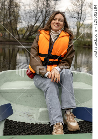 Woman wearing an orange life jacket relaxes in a small boat on a tranquil lake, surrounded by lush trees on a cloudy spring day. 129602194