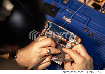 A technician meticulously repairs a smartphone battery, using tools to connect components in a well-organized workshop setting. 129602739