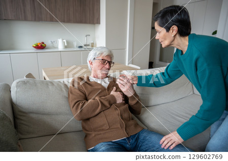 Caring wife helps elderly suffering husband clutching chest sitting on sofa, offering glass of water 129602769