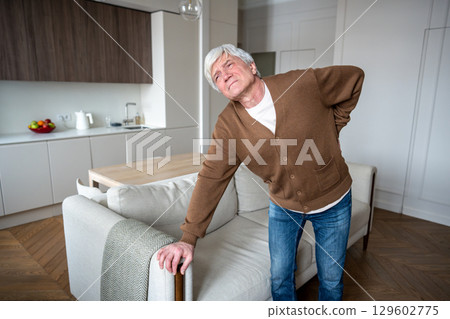 Squinting elderly man suffers alone gripping aching spine with limited mobility in empty house Squinting elderly man suffers alone gripping aching spine with limited mobility in empty house 129602775