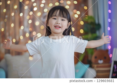 Happy Child in White T-Shirt with Open Arms in Bright Room with String Lights 129603681