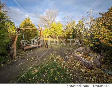 Old country cottage courtyard with a large bench, swing. Large bench swing in garden in autumn Old country cottage courtyard with a large bench, swing. Large bench swing in garden in autumn 129604618