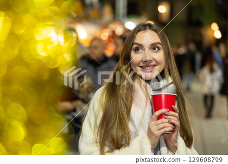 Young woman stands with glass of hot coffee in her hands and enjoys festive New Year atmosphere Young woman stands with glass of hot coffee in her hands and enjoys festive New Year atmosphere 129608799