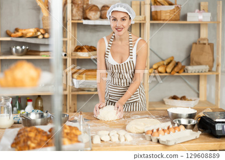 Female baker kneading dough on floured table in bakery 129608889