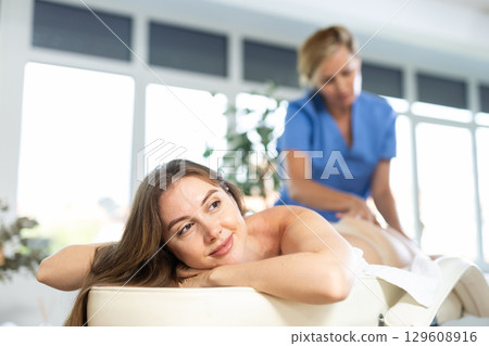 Relaxed young woman lying on massage table enjoying remedial body massage done by professional masseur in modern spa salon 129608916