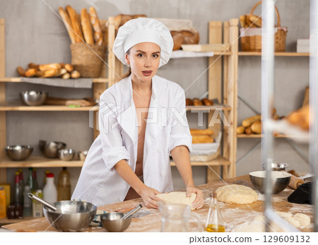 Smiling relaxed young sexy girl professional baker topless in white uniform standing at work table and kneading dough during working day in bakery 129609132
