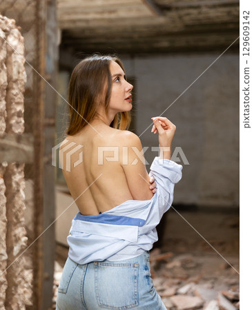 Rear view of woman standing in shirt with bare back inside destroyed building 129609142