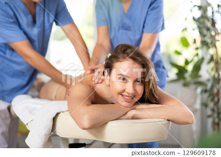 Relaxed young woman having body massage therapy conducted by two masseurs 129609178