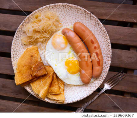 Fried sausages with scrambled eggs, pancakes and cabbage on a wooden background Fried sausages with scrambled eggs, pancakes and cabbage on a wooden background 129609367