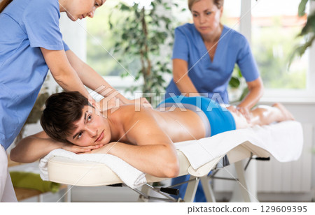 Relaxed young man having back massage therapy conducted by two masseuses Relaxed young man having back massage therapy conducted by two masseuses 129609395