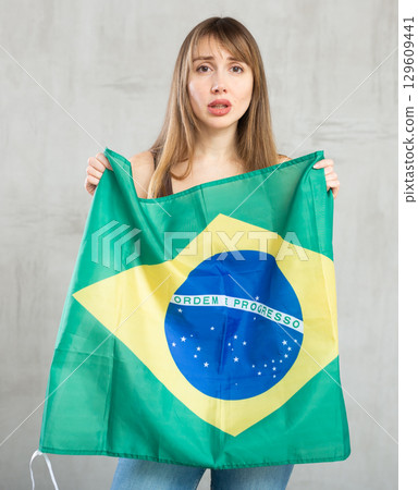 Sad young woman holding flag of Brazil against unicoloured background 129609441