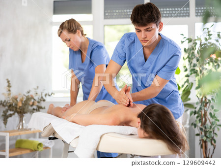 Two female and male masseuses perform stroking and rubbing of muscles of back and shoulders to girl 129609442