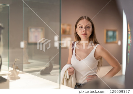 Young female visitor stands near glass display case and examines valuable exhibit Young female visitor stands near glass display case and examines valuable exhibit 129609557