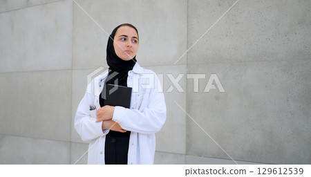 Medical professional wearing hijab and white coat standing near gray wall, holding medical textbook with pensive expression, reflecting healthcare knowledge and deep thinking Medical professional wearing hijab and white coat standing near gray wall, holding medical textbook with pensive expression, reflecting healthcare knowledge and deep thinking 129612539