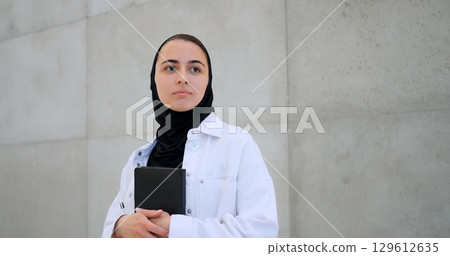 Muslim woman wearing hijab, white shirt, holding book close, gazing pensively near concrete wall, embodying contemplative moment 129612635