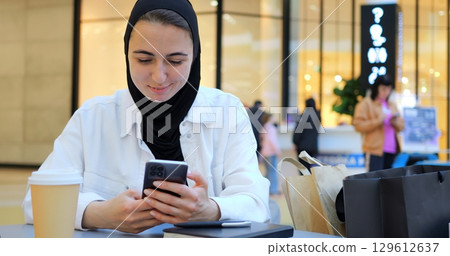 Young muslim woman wearing hijab sitting at table in shopping mall, comfortably browsing smartphone with relaxed smile, embodying modern digital lifestyle and urban connectivity 129612637