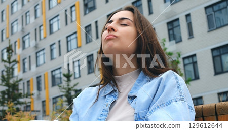 Confident young woman with long brown hair, wearing a light blue denim shirt over a beige top, poses and claps her hands in front of a modern residential building with gray and yellow facade Confident young woman with long brown hair, wearing a light blue denim shirt over a beige top, poses and claps her hands in front of a modern residential building with gray and yellow facade 129612644