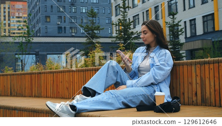 Comfortable woman relaxing on wooden bench, browsing smartphone with takeaway coffee cup nearby, enjoying peaceful moment during urban day Comfortable woman relaxing on wooden bench, browsing smartphone with takeaway coffee cup nearby, enjoying peaceful moment during urban day 129612646