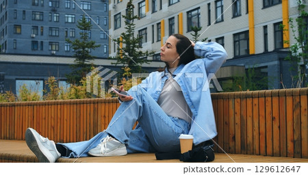 Young woman wearing casual clothes sits on a wooden bench, sipping coffee from a disposable cup and browsing her smartphone, against the backdrop of modern buildings and greenery Young woman wearing casual clothes sits on a wooden bench, sipping coffee from a disposable cup and browsing her smartphone, against the backdrop of modern buildings and greenery 129612647