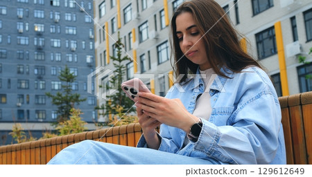 Young woman wearing casual denim jacket sitting on urban bench, browsing internet and engaging with mobile app while enjoying peaceful moment in modern city environment Young woman wearing casual denim jacket sitting on urban bench, browsing internet and engaging with mobile app while enjoying peaceful moment in modern city environment 129612649