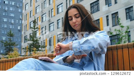 Sitting on a bench, a young woman wearing casual clothes checks the time on her smartwatch while holding a smartphone, possibly waiting for someone or something 129612672