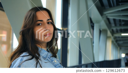 Young woman with long brown hair, wearing a denim shirt, sips coffee from a disposable cup in a modern building with large windows and metal framework Young woman with long brown hair, wearing a denim shirt, sips coffee from a disposable cup in a modern building with large windows and metal framework 129612673